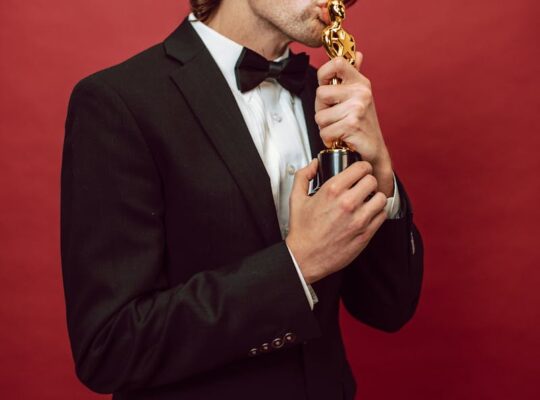Man in a tuxedo kissing a trophy, symbolizing achievement, against a vibrant red background.