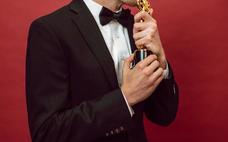 Man in a tuxedo kissing a trophy, symbolizing achievement, against a vibrant red background.