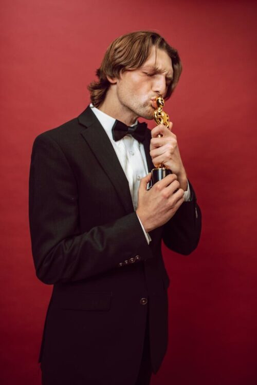 Man in a tuxedo kissing a trophy, symbolizing achievement, against a vibrant red background.